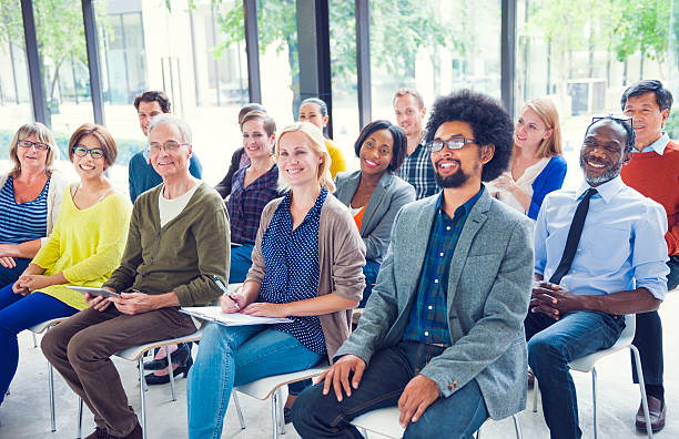 A diverse group of adults sitting in rows of chairs, attentively listening and smiling in a well-lit indoor setting with large windows.