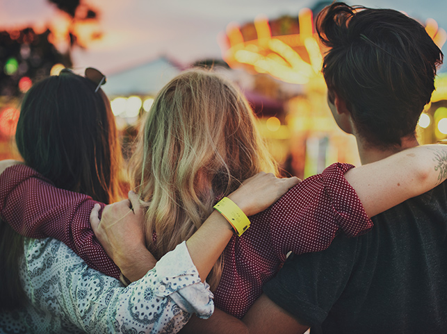 Three friends with their arms around each other, looking out at a carnival or fair during sunset.