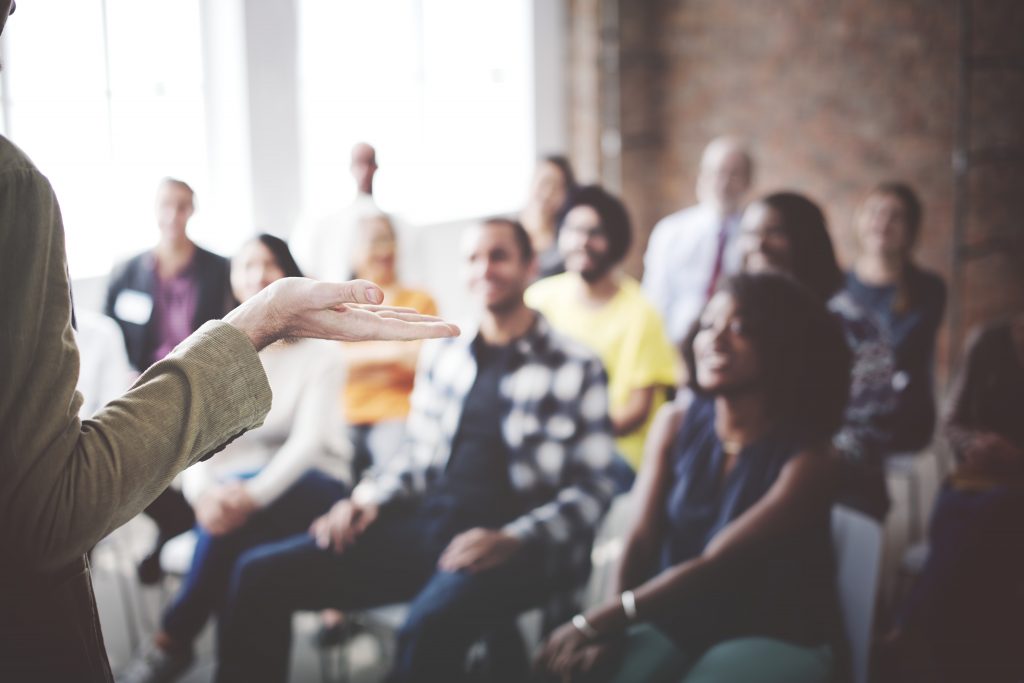 A close-up of a speaker's hand gesturing while addressing a group of attentive listeners.