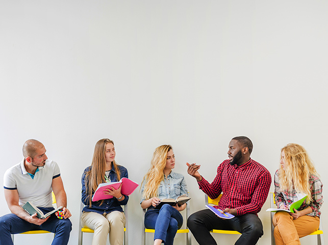 A diverse group of five people sitting in a semi-circle, engaged in a discussion, with some holding books and notes.