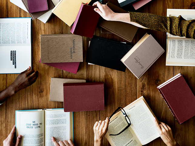 Overhead view of several hands reaching for and holding various books on a wooden table.