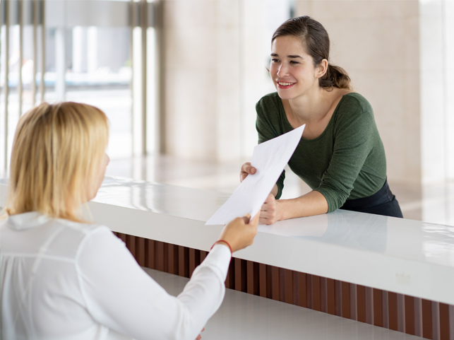 A young woman leaning over a counter, smiling and talking to another woman show a WISE description document.