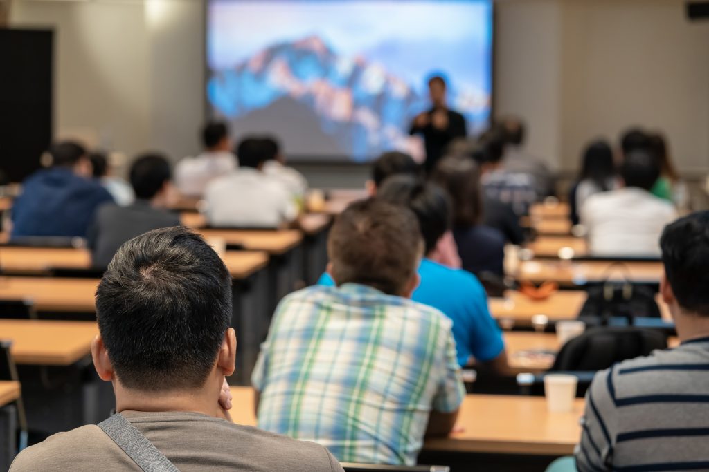 A diverse group in a lecture hall attentively listening to a presenter standing in front of a projection screen