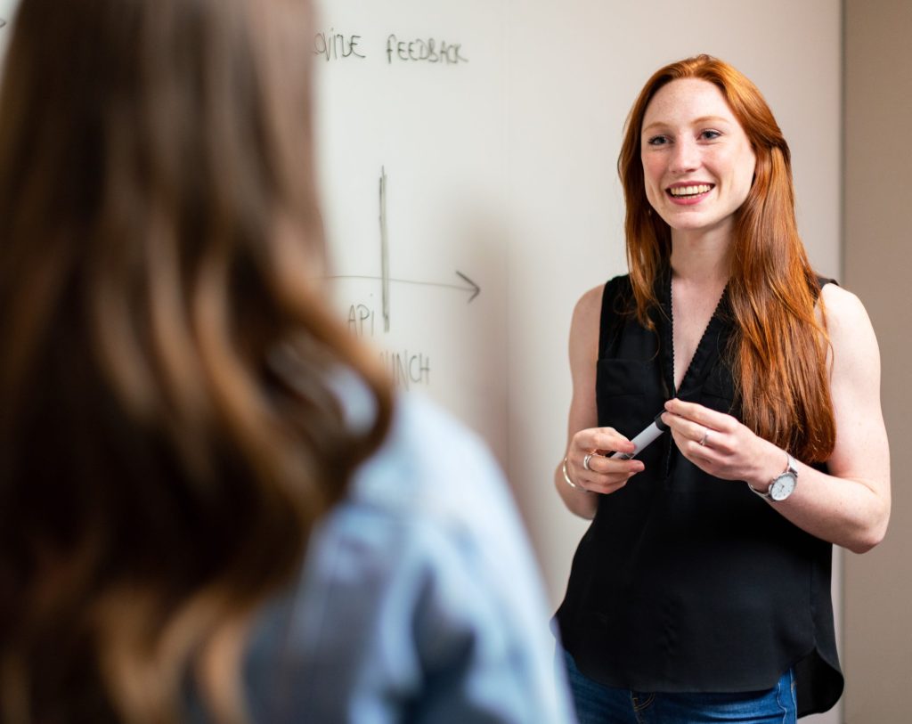 A woman with long red hair presenting in front of a whiteboard.